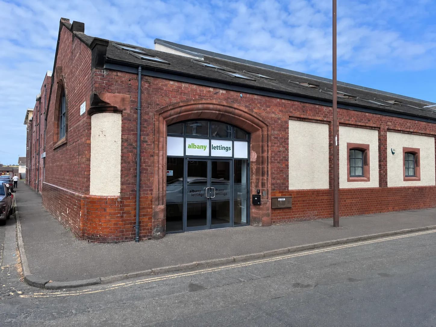 A red brick building with an arched entrance housing Albany Lettings, located on a street corner with a paved sidewalk and asphalt road.