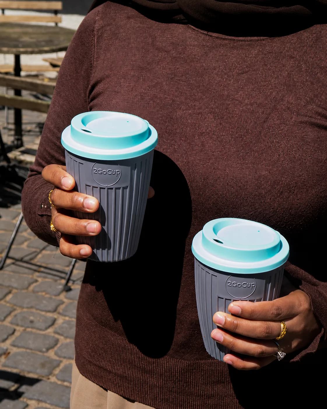 A person holds two gray reusable coffee cups with light blue lids, one slightly closer and larger than the other, against a background of a cobblestone street and blurred outdoor furniture.
