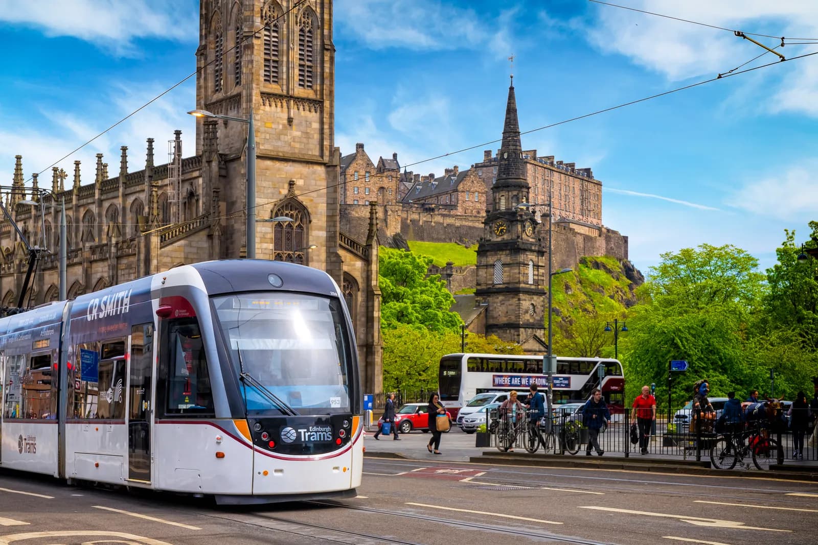A modern tram with "Edinburgh Trams" visible on its front travels on a street with historic stone buildings and a castle on a hill in the background under a blue sky.