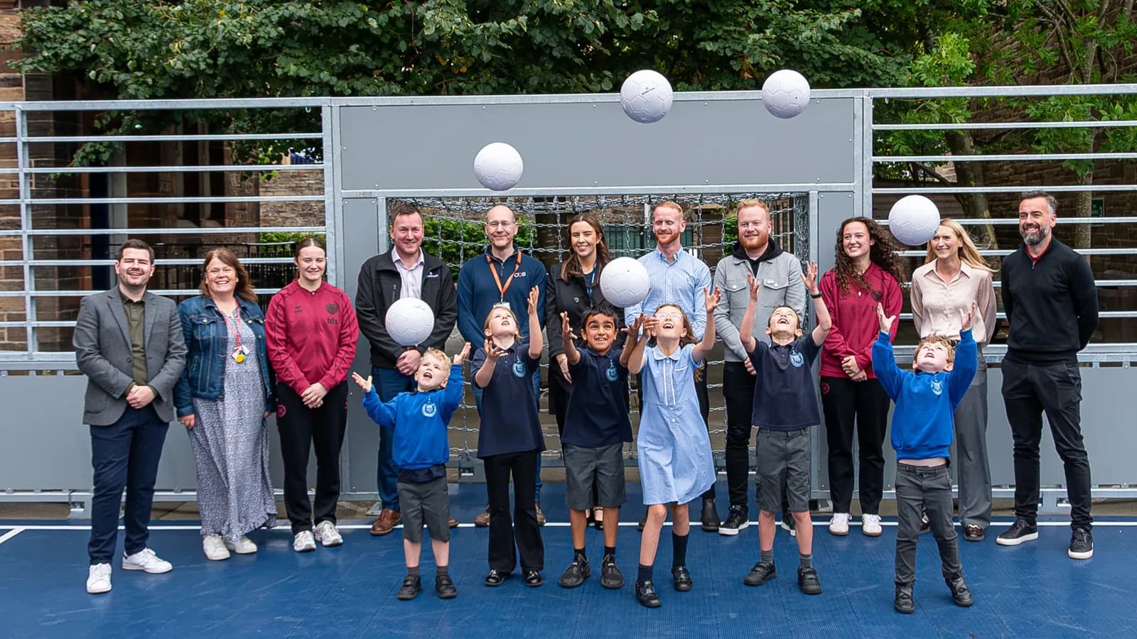 A group of adults and children stand on a blue court with soccer balls in the air, in front of a goal with trees behind it.